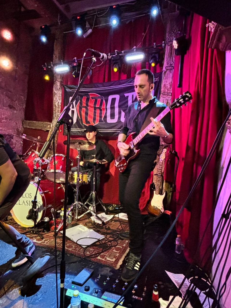 Yor tocando una guitarra roja SG en la sala Los Clásicos con el batería al fondo
